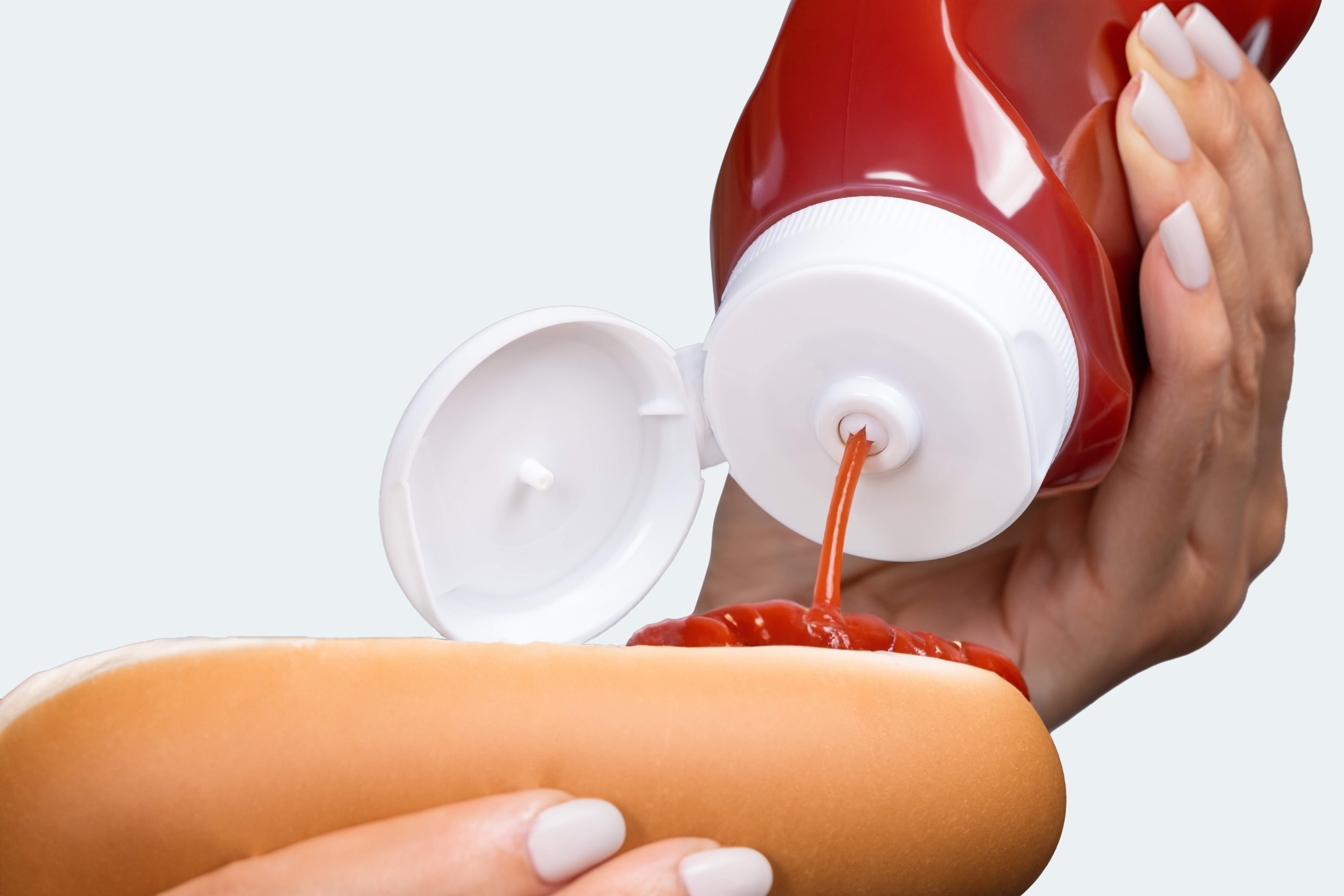 A person squeezing kethcup from an inverted ketchup bottle onto a hot dog